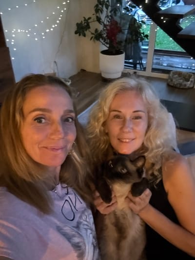 Two women are taking a selfie with a cat. The woman on the left has long blonde hair and is wearing a light purple t-shirt with a graphic print. The woman on the right has curly blonde hair and is wearing a black top. She is holding a Siamese cat. The background shows an indoor setting with string lights, a potted plant, and a staircase. The location is identified as Steklyannyi, Russia.