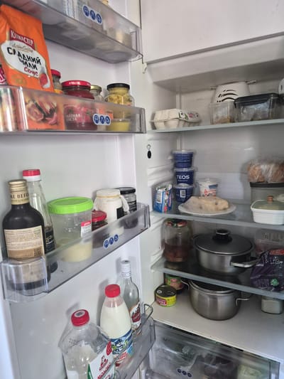 The image shows the interior of a refrigerator. The refrigerator is filled with various food items, including jars of sauces and condiments, containers of dairy products, eggs, a cake, and pots. There are also bottles of liquids, such as oil and milk. The refrigerator appears to be organized, with items placed on shelves and in drawers. The lighting in the refrigerator is standard for such appliances. The image is taken from a slightly elevated angle, looking down into the open refrigerator. The background is a white interior wall of the refrigerator.