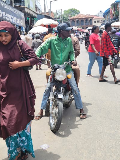 The image depicts a bustling street scene in Suleja, Nigeria. A man in a green jacket and jeans is sitting on a red and silver motorcycle in the middle of the road. A woman in a maroon hijab and a patterned dress is standing nearby. Other people are visible in the background, including individuals wearing casual clothing such as a red t-shirt and a red plaid shirt. The environment shows various shops and buildings along the street, with many stalls and umbrellas indicating market activity. The street is paved, and the overall impression is a crowded, active urban environment.