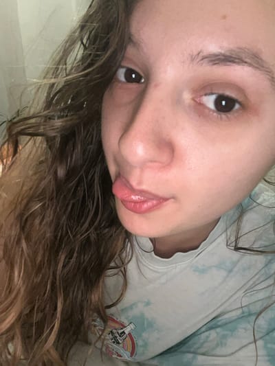 A selfie taken by a young woman with light skin, dark eyes, and brown hair. She is wearing a tie-dye t-shirt with a Coca-Cola logo and rainbow graphic. The photograph is rotated, with the woman's face angled towards the upper left of the frame. Her hair is partially obscuring the lower portion of the image. There is no indication of the location being Saarbrücken, Germany.