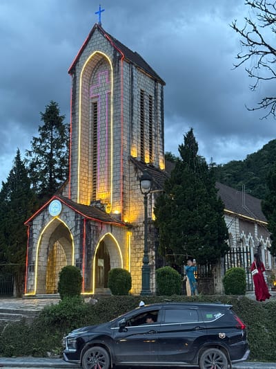 The image shows the exterior of the Stone Church in Sa Pa Ward, Vietnam, decorated with string lights and a neon cross. The church is constructed of stone, with arches and a tiled roof. A dark-colored SUV is parked on the right side of the frame. People, including one in a blue dress taking a picture, are visible near the church. There are trees and greenery around the church. The sky is overcast.
