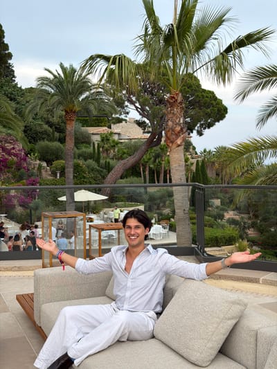 A man wearing a white button-down shirt and white pants is sitting on a light grey couch. He is smiling and has his arms outstretched, palms up, towards the viewer. The man is wearing bracelets on his left wrist and a watch on his right wrist. He is wearing dark brown shoes. The couch is placed on a stone patio. In the background, there are palm trees, lush green vegetation, and buildings. There are people dining at tables in the distance, and a white umbrella is visible. The scene appears to be an outdoor dining or event area. The location is identified as Vallauris, France.