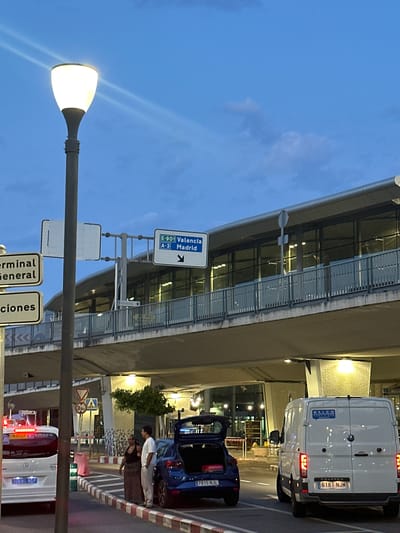This image shows an exterior view of Valencia Airport in Manises, Spain, at dusk. A street lamp with a glowing white light is positioned on the left side of the frame. In the background, the airport terminal building, characterized by its modern architecture with large glass windows and a prominent overhang, is visible. Road signs indicating directions to "Valencia" and "Madrid" are mounted on poles above the roadway.
Several vehicles are present: a white van with the license plate "6181 NJM" is parked on the right side of the road, facing away from the camera. To its left, a dark blue hatchback car with its trunk open is parked, revealing luggage. A white taxi is also visible on the far left, with its red hazard lights illuminated.
Two individuals are standing on the sidewalk next to the blue car. One person is wearing a dark dress and the other is wearing a light-colored shirt and pants. They appear to be interacting. The ground is paved, with a red and white striped curb delineating the roadway from the sidewalk. Traffic signs, including a yield sign and pedestrian crossing signs, are also present. The sky is a deep blue with some scattered clouds, indicating twilight conditions.