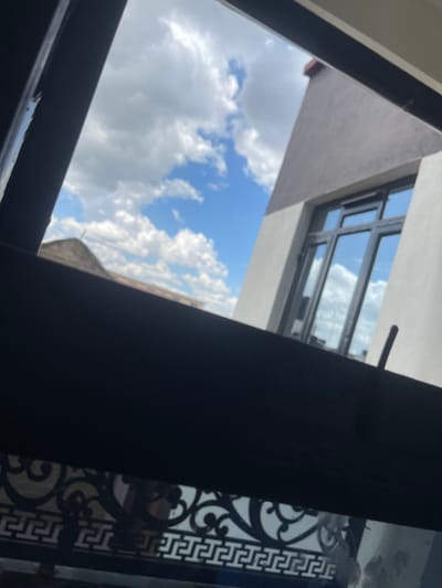 The scene presents an exterior view observed through a shadowed window or opening in Thika, Kenya. The upper two-thirds of the image display a bright blue sky with numerous scattered white cumulus clouds. A modern building occupies the right side of the frame, featuring a light-toned facade, a contrasting darker brown upper section, and a large multi-paned window system reflecting the sky and clouds. Towards the left, the reddish-brown roof peak of an older building is visible against the horizon. A prominent dark horizontal structure, likely part of the immediate foreground's window frame or balcony, bisects the lower portion of the image. Below this, a black wrought-iron railing with ornate scrollwork and a repeating Greek key-like pattern is visible. No individuals are present or engaged in observable activities within the frame.