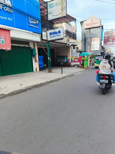 The image shows a street scene in Kecamatan Margahayu, Indonesia. The location is identified by the coordinates: latitude -6.967, longitude 107.576. 

The scene is captured from a moving vehicle, resulting in motion blur. A person is riding a motorcycle towards the camera, while multiple businesses and shops line the street on the right side. The shops are identified by signs including "Mie Baso", "Remaja Studio Foto", "Sari Edibles", and "Kartika Sari". There are several people on foot, one of whom is sitting on a bench in front of a shop. In the background, there are other vehicles and buildings. The pavement is grey and rough, and the sky is cloudy.