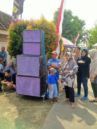 The scene is located in Kecamatan Kedawung, Indonesia, at the coordinates latitude -7.514, longitude 111.03. There is a stack of four large speakers with purple grilles on a paved area in front of a group of people. The speakers are secured with blue straps. A young boy wearing a blue shirt and green cap stands next to the speakers. To his right is a man in a blue and white striped shirt, with his arms folded across his chest.  A woman wearing a gray headscarf and patterned dress stands to the right of the man, her hands clasped in front of her. A woman wearing a black shirt with a white emblem on the front and black pants stands to the right of the woman in the gray headscarf. A woman wearing a white headscarf with a black dress stands to the right of the woman in the black shirt. A man wearing a red shirt and khaki pants stands to the right of the woman in the white headscarf. Two children stand in the background, one on each side of the speaker stack. A man in a dark blue shirt stands behind the children.  In the background, there is a red and white flag, greenery, and a white tent.