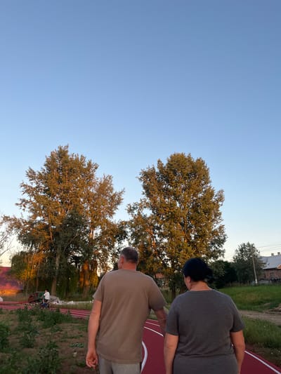 Two individuals, a male and a female, are walking away from the camera on a red running track. The male is wearing a brown t-shirt and grey pants, while the female is wearing a dark grey t-shirt and a dark head covering. They are holding hands. In the background, there are several large trees with autumnal foliage, illuminated by the setting sun. A young boy is riding a bicycle on a path to the left of the running track. Further in the distance, buildings with red roofs are visible, along with other trees and a grassy area. The sky is clear and blue. The scene appears to be a park or recreational area.