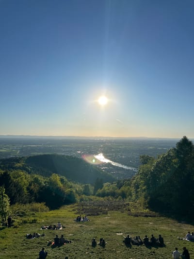 The image depicts a landscape scene in Heidelberg, Germany, likely taken from Philosophenweg (Philosopher's Way). The sky is a clear, gradient blue, brightest near the sun. The sun is centrally placed with sunbeams radiating from it. The cityscape of Heidelberg stretches across the horizon, with the Neckar River visible as a shining strip of light. Forests and greenery cover the hillside, including trees at the edge of the frame and along the river. In the foreground, there is a grassy hill where people are resting, sitting, and socializing. The composition is oriented vertically. The overall impression is one of a sunny day with an elevated perspective over the city.