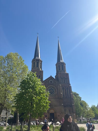 The image shows a low-angle view of the Saint Paul’s Church in Antwerp, Belgium. The church features two prominent spires and is constructed of brown brick. In the background, a clear blue sky is visible, with a contrail of an airplane. The foreground contains green trees that partially obscure the base of the church and some people. The church is located in a bustling environment with bikes parked, indicating the presence of people.