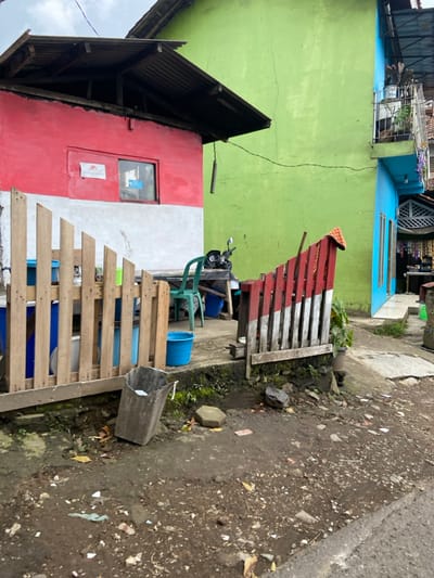 The image shows a view of a street in Lembang, Indonesia, located at coordinates -6.835 latitude and 107.602 longitude. Two adjacent houses are visible, one painted in red, white, and green with a black roof, and the other in light green and blue, with a white roof. Both have wooden fences in front. There is a motorcycle parked outside the red, white, and green house. A wooden fence in front of the red, white, and green house has fallen over, and is propped against the wall. A blue bucket is visible on the ground. The ground is covered with dirt and debris.