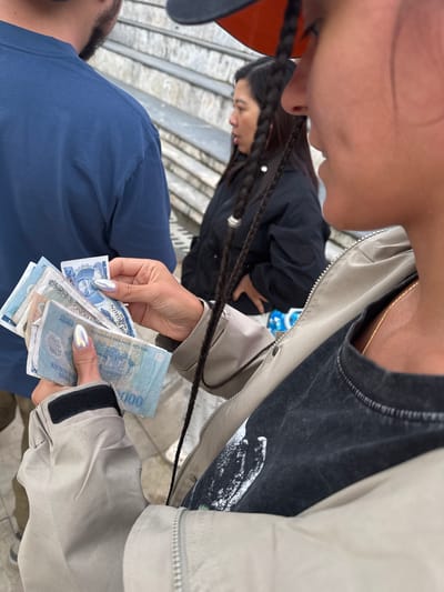 The image shows a close-up, high-angle view of two individuals interacting, likely during a transaction. One person is holding Vietnamese Dong banknotes, including denominations of 100,000 VND. The other person, wearing a blue shirt, appears to be receiving or giving the money. The background is blurred, revealing stairs, and the overall scene suggests an outdoor location. The individual holding the money is wearing a jacket with a beige and black design. While the setting is not definitively confirmed, the presence of Vietnamese Dong currency suggests the location is in Vietnam. If this location is Sa Pa Ward, Vietnam, then this is likely a financial transaction occurring in a public or tourist area within the ward.
