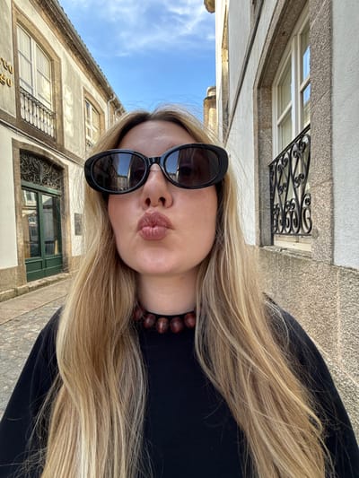 A young woman with long blonde hair and dark sunglasses poses for a selfie in a narrow European-style street, possibly in Santiago de Compostela, Spain, given the architecture of the buildings. The street is paved with cobblestones. The woman is wearing a black long-sleeved shirt and a necklace made of large, dark beads. She is puckering her lips for the camera. The buildings flanking the street are old, with stone facades, arched windows, and decorative iron grates. The sky above is blue with scattered clouds.