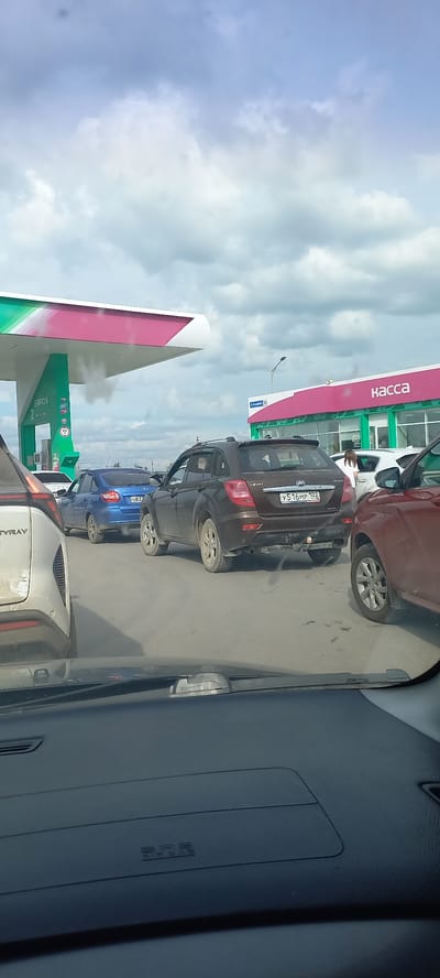 The image shows a view from inside a car, looking out at a gas station in Sibay, Russia. The scene depicts a line of cars waiting at the gas station. The weather appears cloudy, with a mix of blue sky and white clouds visible.

Several cars are parked in a queue. The most prominent vehicles are a dark brown sedan with a license plate reading "У516МР 102", a blue hatchback with a license plate "083", and a red car further down the line. The gas station features a pink and white canopy with green accents. Text visible on the canopy includes the word "КАССА" (KASSA), which means "cash desk" or "checkout" in Russian. Another sign on the gas pump area displays "EUROS" and what appears to be fuel price information. The interior of the car from which the photo is taken is visible, showing a dark dashboard and a reflection of a side mirror. The overall impression is a mundane scene of cars waiting to refuel at a gas station on a cloudy day.
