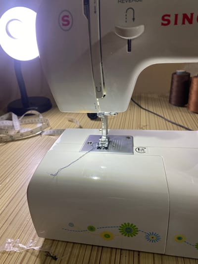 The image shows a close-up, eye-level view of a white Singer sewing machine. The machine is situated on a light-colored, woven mat. In the background, to the left, is a bright ring light attached to a black stand. To the right of the sewing machine, two spools of brown thread are visible. A white measuring tape with black markings is draped across the mat in the mid-left of the frame. A dark gray thread is lying on the sewing machine's needle plate and extending onto the machine's body. The environment does not provide any specific indicators that place the image in Bukhara, Uzbekistan.