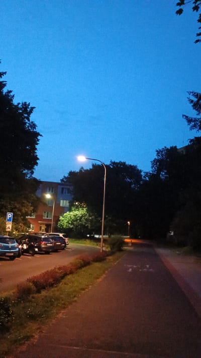 The image depicts a street scene at dusk in Sokolov, Czechia. The sky is a deep blue, indicating twilight. A street lamp illuminates the scene, casting a warm glow on the asphalt road and the surrounding vegetation. Trees line the right side of the road, with a grassy verge between them and the asphalt.
Several cars are parked along the left side of the road, some are dark-colored and appear to be SUVs or sedans, while others are lighter blue. One of the cars has its headlights on. A bicycle is visible leaning against one of the parked cars. In the background, a multi-story apartment building with several windows is visible, some of which are lit from within. Another, smaller street lamp is visible further down the road, its light orange.
There is a blue parking sign with a white "P" symbol and an arrow on the left side of the road. The overall mood of the image is calm and peaceful, suggesting a quiet evening in a residential area. The time of day is clearly dusk or early evening, as evidenced by the bright sky contrasted with artificial lighting. The location is identified as Sokolov, Czechia. There is no discernible weather activity.