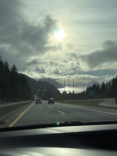 A highway stretches forward, lined with green vegetation and evergreen trees. Power lines and a green "Exit" sign are visible on the shoulder. Cars travel in the same direction. In the distance, mountains rise amidst heavy, low-lying clouds, partially obscuring a body of water reflecting the sunlight. The sun breaks through the clouds, casting a bright glow. The image is taken from inside a vehicle.