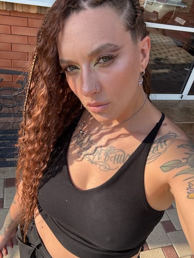 The media file depicts a person with long, curly, reddish-brown hair and distinctive braided elements near the front hairline. They are wearing a black, ribbed tank top and a silver chain necklace. The person has visible tattoos on their upper chest and arm, as well as a nose ring and an earring. Their makeup includes an application of what appears to be green eyeshadow.
The subject is positioned outdoors, with the warm sunlight casting shadows, suggesting a daytime setting, likely during sunny weather. The background includes brickwork, indicative of an outdoor patio or walkway, and a blurred building facade. The image is taken at an upward angle, focusing on the upper body and face of the person. No text is visible in the image. The overall impression is one of casual outdoor activity in a moderately urban or suburban environment.