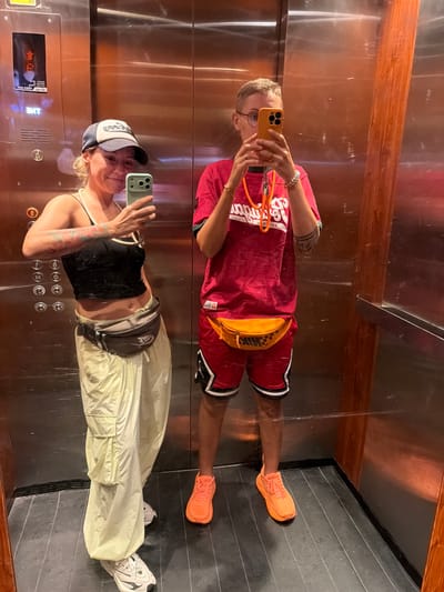 Two individuals are taking a mirrored selfie in an elevator. The reflection shows a person on the left wearing a red jersey with a Nike fanny pack and orange sneakers. On the right, a person wearing a black tank top, light-colored cargo pants, a baseball cap, and white sneakers is also visible. Both are holding up their phones to capture the reflection. The elevator walls are metallic with wooden accents at the top. The floor of the elevator appears to be dark grey and tiled. The image is well-lit, suggesting it was taken indoors during the daytime. The overall mood seems casual and playful. Text on the fanny pack appears to read "NIKE".