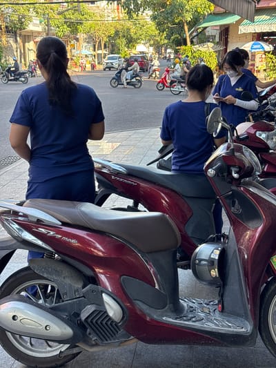 The image captures a street scene in South Quy Nhon Ward, Vietnam, likely during daytime with ample sunlight creating shadows. In the foreground, two individuals wearing dark blue uniforms are partially visible, with their backs to the camera. They appear to be standing on a sidewalk next to several parked motorcycles. The motorcycles are dark red and appear to be scooters, with one having a "VISION" logo visible on its side panel. Another motorcycle has an "SH mode" logo. A black helmet with a carbon fiber pattern is hanging from one of the motorcycles.
In the background, the street is moderately busy with vehicular traffic, including motorcycles and cars. Several people are seen on motorcycles, some wearing helmets and masks. Buildings line the street, with awnings and greenery suggesting a commercial or residential area. The overall impression is one of everyday urban life in a Vietnamese city.