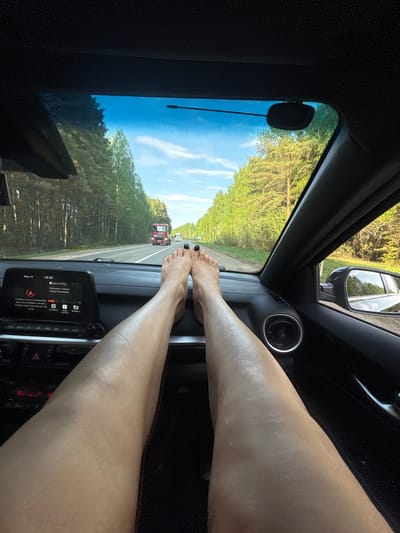 The image shows an interior view from the driver's seat of a car traveling down a road flanked by trees, presumably in a rural area. A portion of the windshield and side mirror are visible, revealing a blue sky and greenery. A truck is visible on the road ahead.

The distinguishing feature of the image is a pair of legs and feet placed on the dashboard of the vehicle, obscuring the car's infotainment system and parts of the interior. The toenails are painted black. 

There is text indicating that the image was taken on May 16th at 19:20. It indicates that a SD card is incompatible. The image does not contain any details that are specific to Uspenskii, Russia.