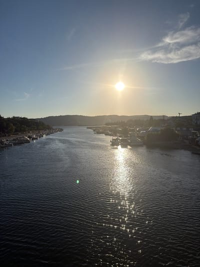 The image captures a view of the Çanakkale Strait in Çanakkale Merkez, Turkey, during a sunny day. The sun is positioned in the upper right quadrant of the sky, casting a bright glare on the water. The strait is calm with gentle ripples reflecting the sunlight, creating a shimmering effect. On the left side of the strait, a line of moored boats and yachts is visible along the shore, with trees and buildings in the background. The right side of the strait also shows moored boats and buildings, with a slightly more developed shoreline. In the distance, rolling hills form the horizon, partially obscured by the sunlight and atmospheric haze. The sky is clear with a few wispy clouds. There are no visible people in the image.
