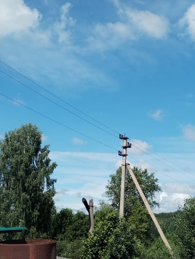 The image depicts an outdoor scene with a bright blue sky filled with scattered white clouds. In the foreground, to the left, is a large, leafy green tree. Dominating the center-right of the frame is a concrete utility pole with multiple insulators and wires running horizontally across the image. A second, diagonal pole supports the main utility pole. Below the utility poles and to the left, a portion of a rusty, cylindrical object, possibly a tank or barrel, with a textured surface is visible. The background consists of dense green trees and foliage, suggesting a rural or semi-rural environment. The overall impression is a clear, sunny day with overhead power lines intersecting a natural landscape. The location is stated to be in the village of Зипуново, Russia.