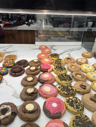 The image displays a vibrant assortment of numerous cookies meticulously arranged on a white, marble-patterned counter, likely within a bakery or sweet shop in Marousi, Greece. On the far left, a tall, vertical glass display case reflects various dark, utilitarian objects, possibly kitchen appliances, as well as some white cups and a dark wooden surface.
The majority of the frame is filled with rows of diverse cookies. In the upper sections, several light-colored, possibly ring-shaped or donut-like cookies with central indentations are visible. Below them are multiple rows of yellowish cookies, uniformly covered with dark chocolate sprinkles.
Further down, there's a prominent collection of pink and reddish-toned cookies. Some are plain and oval, while others are round, featuring decorative swirls or contrasting centers, often with a vibrant pink or white frosting. Many of these circular cookies exhibit a two-toned base, combining pink with chocolate brown, with a distinct central swirl.
The lower part of the display predominantly features darker, chocolate-based cookies. Many have a visible white or light-colored filling at their center. Other chocolate cookies are topped with colorful candy-coated chocolate pieces, resembling M&Ms, or chunks of dark chocolate. One cookie at the bottom right distinctively incorporates an Oreo-like pattern.
The cookies are brightly illuminated, emphasizing their varied textures, colors, and decorations. The arrangement suggests a retail presentation of baked goods. There are no people or visible text in the image.