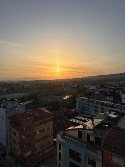 The image shows a sunset over what appears to be a cityscape. The sun is low on the horizon, creating a warm, golden glow. In the foreground, there are buildings of various styles and heights, mostly rooftops, including some with red tiles. Below the buildings is a green area that appears to be the city center. In the distance, there are mountains. The image orientation is sideways. It is not possible to determine if this is Oviedo/Uviéu, Spain.