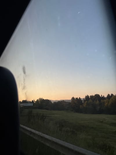 The image shows a rural landscape, likely in the vicinity of Kasak, Bulgaria, viewed from inside a vehicle. The sky transitions from a light blue at the top to a soft orange and yellow hue near the horizon, indicating either sunrise or sunset. A dense line of trees forms a dark silhouette against the lighter sky, with a few taller pine trees interspersed among them. In the foreground, a grassy field stretches out, appearing somewhat unkempt with taller weeds. A white guardrail runs along the bottom edge of the frame, suggesting a road. In the distance, on a slightly elevated area behind the trees, a white truck trailer is visible, possibly part of a larger vehicle or structure, with some dark smoke rising from behind it. The overall impression is of a tranquil, natural setting observed during twilight hours.