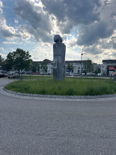 This is a photograph of a stone sculpture in a park-like setting. The sculpture is abstract, appearing to be two figures back-to-back or leaning against each other. It is made of rough, gray stone. The sculpture is positioned on a patch of grass next to a paved pathway. In the background, there are modern buildings with windows, trees, and cars parked along the side of the road. The sky is dramatic, with a mix of dark, heavy clouds and bright patches where the sun is shining through, suggesting a dynamic weather system. The image is oriented vertically, with the sky occupying the top half and the ground the bottom.

Notable details include the distinct clouds, indicative of possibly late afternoon or an approaching storm, and the modern architecture in the background. The overall mood is somewhat somber due to the clouds, but the bright sunlit areas offer a sense of hope. There is visible text on a billboard in the background that reads "NR.36 SCHARF."