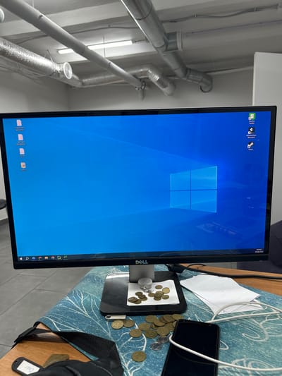 The image shows a computer monitor displaying the Windows 10 operating system. The desktop has several icons, including "Bambu Studio," "Wondershare Recoverit," and "Steam." On the left side of the screen, there are three text files labeled in Russian: "код для все врачи.txt," "текст для рекламы Д...," and "c12.txt," along with an icon labeled "bot v1." The monitor is a Dell brand.

In front of the monitor, on a desk covered with a blue and white patterned mat, are several objects. These include a pile of coins, some loose coins scattered around, a smartphone with a white charging cable, a black bag strap with a tag that says "Chef," a USB flash drive, and a stack of white paper. The background shows industrial-style piping and lighting, typical of an office or workshop environment. The date and time displayed on the monitor's taskbar indicate 9:50:21 on June 29, 2026. This scene is indoors. The image does not provide specific details to locate it in Kamianske, Ukraine, beyond what is stated in the prompt.