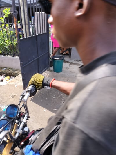 The image depicts a street scene in Alaiyabiagba, Nigeria, captured from the perspective of an individual seated on a motorcycle. The foreground features the right side of a dark-skinned individual's head, shoulder, and arm. This person wears a dark shirt and a dark cap. Their right hand, adorned with a yellow and black glove, grips the chrome handlebar of a motorcycle, which displays blue-faced gauges. A red bracelet is visible on the individual's right wrist. A black bag and a blue cloth are visible on the motorcycle's tank area.
In the midground, a dark grey metal gate, partially ajar, reveals an entrance. To the left of the gate, green foliage grows, and the ground is littered with debris including white paper and plastic. Beyond the gate, in the background, a second individual is partially visible, wearing a bright pink garment and a patterned lower garment (likely a skirt or wrapper), standing inside what appears to be a paved entrance. A dark green bucket containing a white substance rests on the ground near this person. The overall environment suggests an urban or semi-urban setting with a mix of natural elements and built structures.