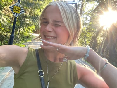 A young woman with blonde hair and a nose ring is taking a selfie in a park. She is wearing a green tank top, a silver necklace, and several rings on her hand. She is winking and smiling, with her arm raised to cover part of her face. In the background, there are trees, sunlight filtering through the leaves, and a "SLOW ZONE" sign. The location is identified as Whistler, Canada.