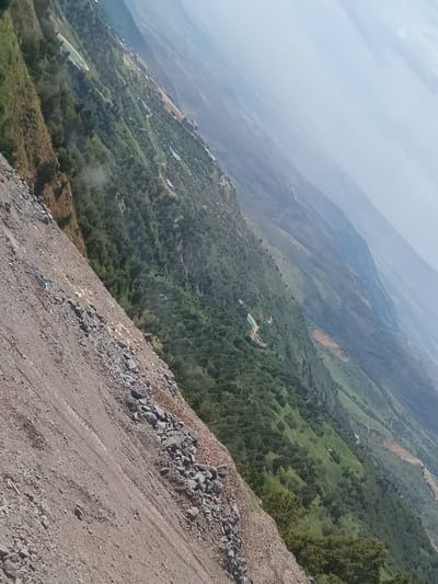 The image depicts a steep, rocky slope in the foreground, transitioning into a densely vegetated mountainside with scattered structures and a distant, hazy mountainous landscape. The vegetation consists of numerous trees and shrubs, forming a green carpet down the slopes. A few buildings, appearing to be houses or small structures, are visible nestled within the trees and along the contours of the mountain. Further into the distance, the landscape softens into rolling hills and plains shrouded in atmospheric perspective, indicating a vast, open environment. The sky is overcast with a light haze, contributing to the diffused lighting. The overall scene suggests a rural or semi-rural mountainous region. Without further context, it is impossible to confirm the specific location within Nurekata, Uzbekistan, as the image lacks any distinct landmarks or identifying features that would definitively place it there. However, the topography and vegetation are consistent with mountainous terrain found in parts of Uzbekistan.