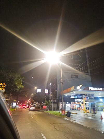 The image depicts a nighttime scene in what appears to be the city of Miraflores, Peru. A brightly illuminated street light is prominently featured, creating a starburst effect. The street is lined with commercial establishments, including a Primax gas station and a Listo! Gourmet convenience store. There are other businesses with signage that say "Led", one reading "DIGITAL OUTDOORS" with a phone number. A price sign for the gas station is in the bottom left.

A line of vehicles, with their headlights and taillights visible, stretches into the distance along the road. The overall environment suggests an urban or suburban setting during the night.