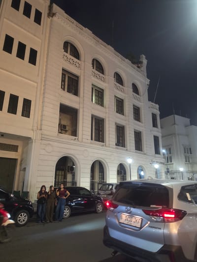 In the city of Medan, Indonesia, at night, three young women stand on a sidewalk next to parked cars. The woman on the left wears a black top and blue jeans. The middle woman is dressed in brown overalls. The woman on the right sports a maroon crop top and blue jeans. They are positioned in front of a multi-story white colonial-style building with arched windows. Across the street, streetlights illuminate the scene, casting long shadows. Several cars are visible, including a silver SUV in the foreground, a black car to the left, and a darker sedan to the right of the women. The building's facade features decorative elements and visible air conditioning units in some of the lower archways. The dark sky suggests it is late evening or night.