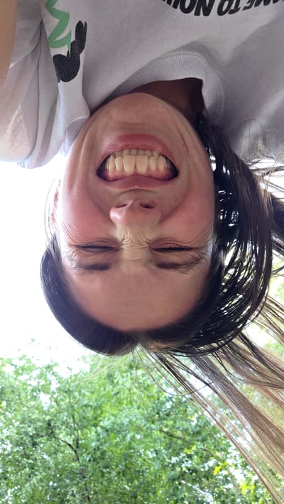 A low-angle, upside-down, close-up shot captures a person's face with their eyes squeezed shut and mouth open in a wide grin, revealing their teeth. The person is wearing a white sweatshirt with a black and green graphic on the left shoulder. Their dark brown hair is splayed out around their head. The background is dominated by bright sky and the green foliage of trees. The scene is outdoors.