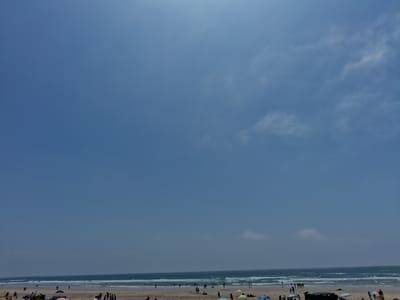 The image shows a beach scene in Figueira da Foz, Portugal. A wide sandy beach is visible at the bottom, occupied by people. The Atlantic Ocean stretches across the middle, with waves breaking near the shore. The sky dominates the upper portion, with shades of blue and scattered clouds. People are walking along the beach and some have set up umbrellas.