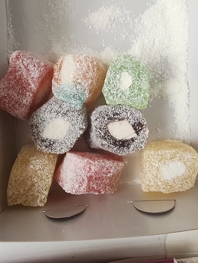 A close-up overhead shot shows a variety of colorful Turkish delight candies, also known as lokum, nestled in a light grey box dusted with white shredded coconut. The candies are square or rectangular in shape and are coated in shredded coconut. Some of the candies are a pale yellow, others are pink, green, or a dark reddish-brown with a white center. One candy in the upper right corner is a creamy white, also covered in coconut. The lighting is soft and even, suggesting an indoor setting. The overall impression is one of a sweet treat, perhaps a gift or a snack. No text is visible in the image. The setting of Ankara, Turkey is provided as context, suggesting these are traditional Turkish confections.