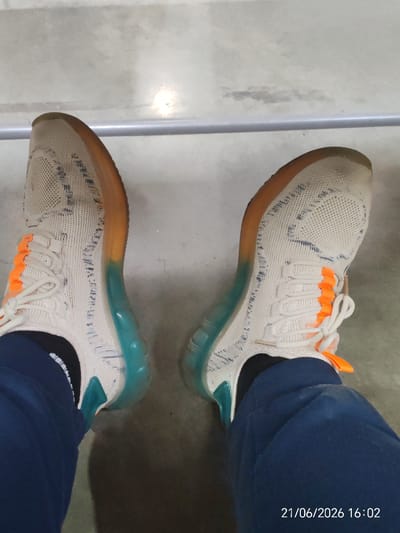 A pair of white and orange athletic shoes with translucent blue and orange soles are shown from a first-person perspective, with the wearer's navy blue sweatpants visible. The shoes appear to be a knit material with a patterned design. The background is a concrete floor, and a metal railing is visible in the upper portion of the image. A timestamp in the lower right corner indicates "21/06/2026 16:02". The scene is consistent with an indoor or outdoor urban environment, possibly a public space such as a park or sports facility in Prikubanskii, Russia.