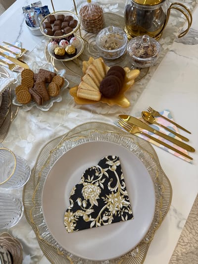 The image depicts a table setting. The table is covered with a pale tablecloth with a floral pattern. A table runner runs along the center of the table. On the left side of the table runner, there are several clear glass containers filled with nuts and candy, as well as small golden bowls filled with an assortment of cookies and chocolates. On the right side of the table runner, there is a place setting with a decorative charger plate, a light gray dinner plate, and a black napkin with gold floral designs. To the upper left of the place setting, there is gold flatware on top of a pastel-colored napkin. Near the bottom of the image, there are a few clear glasses. The environment appears to be indoors. There is no specific information available to indicate the location is in Nottingham, United Kingdom. No people are visible.