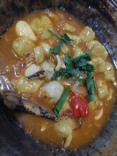 A close-up, overhead shot captures a hearty stew in a rustic, dark-colored pot. The stew features golden, irregularly shaped chunks, likely potatoes or dumplings, submerged in a rich, orange-hued broth. Several ingredients are visible: sliced garlic cloves, slivers of red onion, and a bright red chili pepper. Glistening fish pieces, with visible scales and dark charred edges, are scattered throughout. Verdant sprigs of cilantro or parsley and sliced green onions add a touch of freshness. The pot's textured surface suggests it is made of cast iron or a similar material. The image is well-lit, highlighting the glossy texture of the stew. The specific location in Jelutung, Indonesia, is not evident from the visual information.