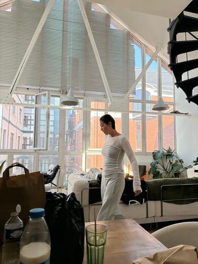 The scene is an interior view of a residential loft or apartment in Saint-Denis, France, characterized by extensive natural light and an industrial-chic aesthetic.

A female individual, appearing to be in her 20s-40s, stands in the mid-ground, facing left in profile with her gaze directed downwards. She has dark hair pulled back and is dressed in a light-colored, long-sleeved, form-fitting top with visible seams, paired with light grey sweatpants.

The room features large, multi-pane windows that extend to a high, sloped ceiling, incorporating white structural beams forming an A-frame. Horizontal blinds cover the upper window sections. A black spiral staircase with metal steps is visible on the far right. A white horizontal bar suspended from the ceiling supports two conical white pendant lights. Behind the individual, a modern sofa with light-colored upholstery and metal frames is present, with a clear plastic bag and other items resting on it. A large potted plant with broad green leaves is positioned behind the sofa on the right.

In the foreground, a wooden table occupies the lower portion of the frame. On the table, from left to right, are a clear plastic bottle of hand sanitizer, a clear plastic bottle containing a white liquid with a light blue cap, a black plastic bag, and a green ribbed glass containing a stirring stick. A brown paper bag stands upright to the left of the individual.

Through the windows, an urban environment is visible under bright, clear daylight conditions. The exterior view includes buildings with reddish-brown brick facades, white window frames, lighter-colored buildings, and a distant, taller structure with a reflective facade.