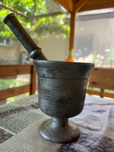 A metal mortar and pestle are placed on a table outdoors, possibly on a porch or patio. The mortar is cylindrical with a flared rim and a pedestal base, decorated with horizontal ribbed bands. The pestle is a simple, elongated cylinder with a slightly flared end. In the background, wooden furniture and lush green foliage are visible, suggesting a natural or garden setting. The overall lighting suggests it is daytime, possibly late morning or early afternoon, with sunlight filtering through the leaves. The scene is set in Karabunar, Bulgaria, implying a rural or suburban location with a focus on a traditional kitchen or herbal preparation tool.