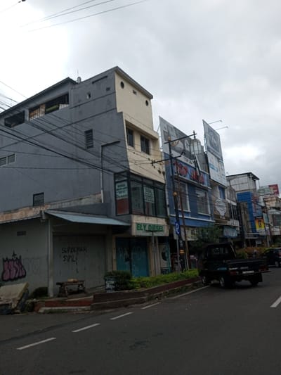 This image depicts a street scene in the city of Vaatharawattai, Sri Lanka, under an overcast sky. The view is of a street, looking along a two-lane asphalt road with a double-dashed white centerline. On the right side of the street, a row of multi-story commercial buildings is visible.
The leftmost building is a three-story structure predominantly grey. Its ground floor features a closed shop with a corrugated metal awning; a rolled-down security door displays white text reading 'SUPREMASI SIPIL'. Pink and purple graffiti, resembling 'JUH', is present on the lower left wall. In front of this shop, a wooden bench or table supports a small, indistinct brown object.
Adjacent to the grey building, another multi-story structure has a cream-colored upper section and a light grey lower section. The ground floor retail space features large glass windows displaying multiple signs with the text 'DIJUAL' (For Sale), and a green fascia sign reads 'ELY DIESEL'. To its immediate right, a closed shop displays light blue graffiti, 'PALADA', on its rolled-down security door.
Further down the street, a blue multi-story building is prominent, adorned with several large advertising billboards on its upper facade, one featuring a stylized 'GO' logo. A tall street pole with an ornate, caged lantern fixture and numerous overhead electrical wires is positioned in front of this blue building. Additional commercial structures with various signs extend into the distance.
A dark-colored pickup truck is parked on the right-hand shoulder of the road, facing away from the viewer. No discernible individuals are present within the visible area.