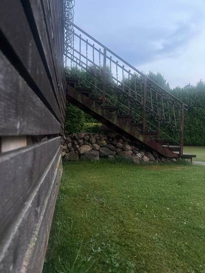 A low-angle outdoor shot shows a weathered, dark brown wooden wall on the left, running horizontally and extending out of frame to the left and bottom. To the right of the wall, a rusty, dark metal staircase with a decorative railing ascends. The staircase is supported by wooden beams and rests on a foundation of large, rounded stones. Beyond the staircase, a dense row of tall, dark green coniferous trees forms a backdrop. The ground in front of the trees and under the staircase is covered in green grass, with a scattering of small white flowers. The sky above is overcast and pale blue, with wisps of clouds. The scene appears to be in a yard or garden setting in Lapės, Lithuania.