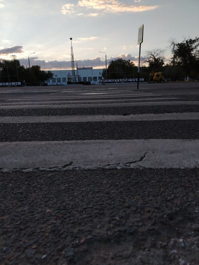 The image shows a pedestrian crossing in Baikonur, Kazakhstan, at dusk. The sky is filled with soft clouds, illuminated by the setting sun, casting a warm glow on the scene. In the background, a white building with large windows stands under a tall structure topped with a star. To the right of the building, a large, yellow and red, spherical sculpture resembling a soccer ball sits with another figure. A yellow pedestrian crossing sign is visible in the middle of the frame, to the right of the pedestrian crossing. The foreground is dominated by the striped pattern of the pedestrian crossing, rendered in dark asphalt and lighter concrete.