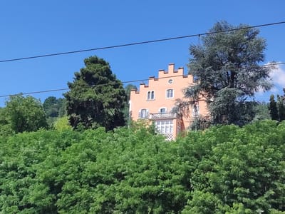 This media file captures a picturesque view of a peach-colored building situated amidst lush greenery under a clear blue sky. The building, possibly a villa or a historical structure, features a distinctive crenellated rooftop and several windows. A balcony with an ornate railing overlooks the dense foliage in the foreground. The scene is dominated by vibrant green trees and bushes, suggesting a natural, possibly mountainous, setting. Two prominent evergreen trees, one to the left and another to the right of the building, add to the landscape's depth. Two dark horizontal lines, likely power or telephone lines, bisect the upper portion of the image, indicating the presence of human infrastructure. The bright sunlight and clear sky suggest it is a pleasant day, likely midday, with fair weather. The absence of people and the overall tranquility of the scene evoke a sense of peace and natural beauty. The location is identified as Brixen - Bressanone, Italy, which is known for its scenic alpine landscapes. No visible text is present in the image.