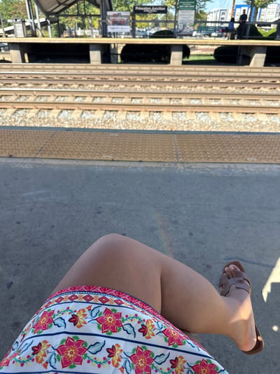 The image depicts a first-person view from a train station platform identified as Princeton Junction, located in West Windsor, United States. In the immediate foreground, the bare legs of an individual are visible, wearing a white skirt or dress with a distinct floral pattern featuring pink, yellow, and orange flowers with blue leaves. The right foot, partially visible, wears a brown open-toed sandal. A subtle, multi-colored light streak is present on the concrete platform surface near the center. Beyond the concrete, a textured yellow tactile warning strip runs along the platform edge. The midground features three visible sets of parallel train tracks, composed of metal rails on wooden ties embedded in light-colored gravel ballast. In the background, across the tracks, an opposing elevated train platform is present, featuring a covered structure supported by concrete pillars. A prominent sign on this far platform clearly reads "Princeton Junction." Several indistinct human figures are observable on the distant platform. Trees with green foliage are visible behind the far platform, and in the furthest background, urban or suburban buildings and vehicles are discernible beneath a clear blue sky. The scene is brightly illuminated by direct sunlight, casting shadows.