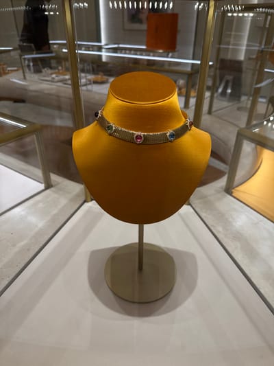 The scene captures a close-up of a luxurious choker necklace displayed on a golden-yellow velvet bust within a glass display case at a retail location in Zapopan, Mexico. The necklace is a gold-toned, articulated band composed of numerous horizontal segments. Seven oval cabochon gemstones are uniformly set along its length, each encircled by smaller, clear accent stones. From left to right, the gemstones appear purple, green, red, pink, light blue, dark blue, and reddish-orange. The bust stands on a gold-colored circular base. The background, out of focus, reveals a high-end retail environment with additional glass and gold-framed display units, linear ceiling lights, and the blurred reflection of a dark-clad individual on the glass. The floor within the display area is light-toned, possibly white or light gray.